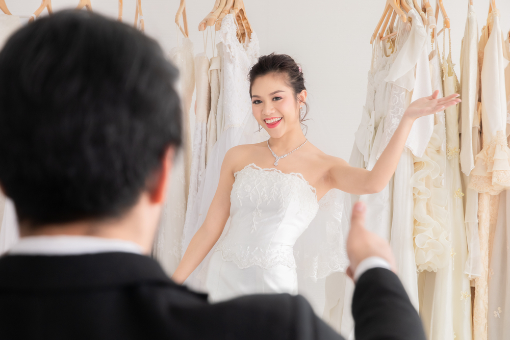 Caucasian groom with beard sitting help bride fitting choosing wedding dress in studio, happy beautiful Asian girl show lace wedding dress to her man